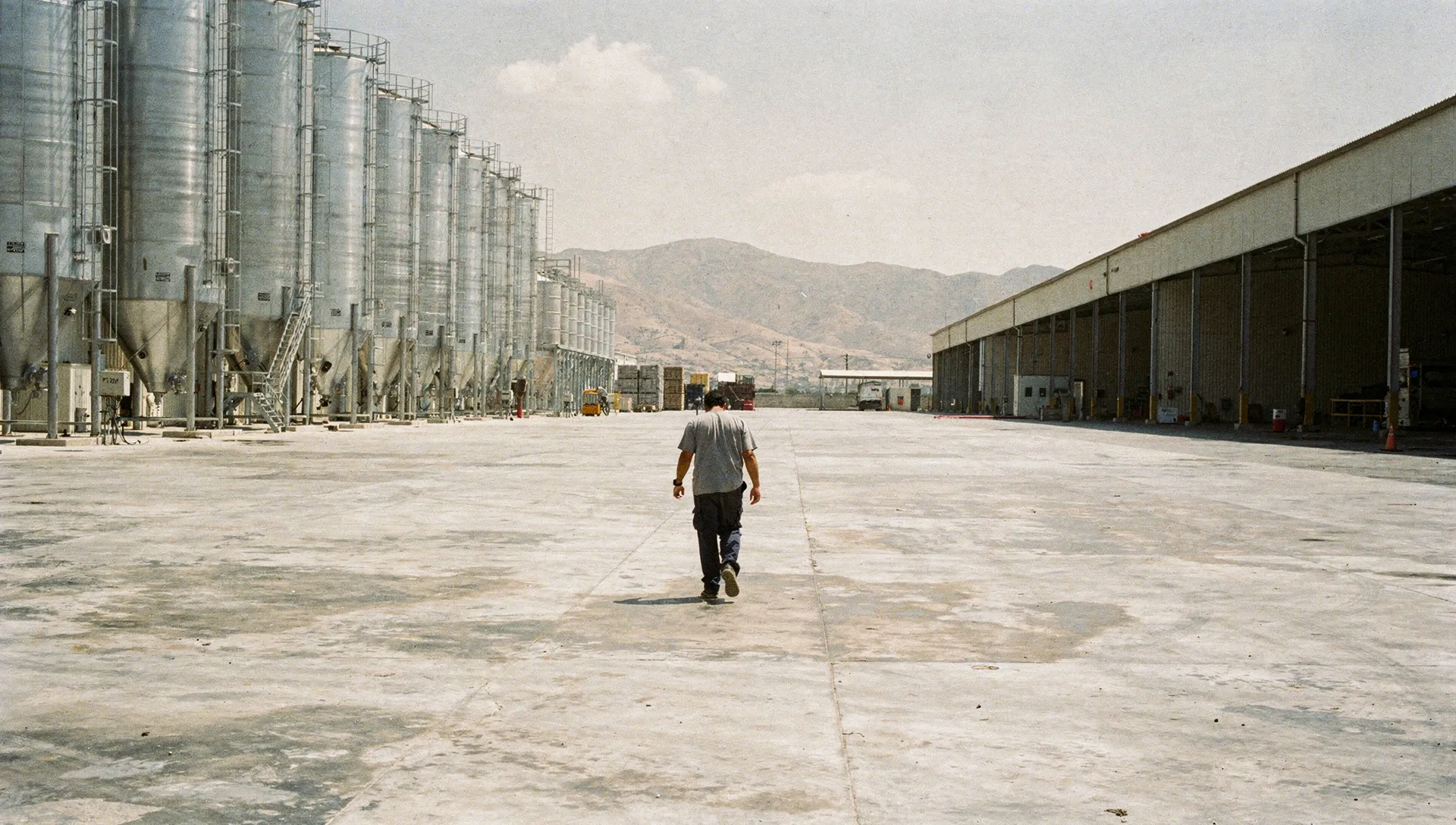 Patio de una planta industrial en el Bajío al mediodía; un trabajador cruza el patio de espaldas entre silos de acero.
